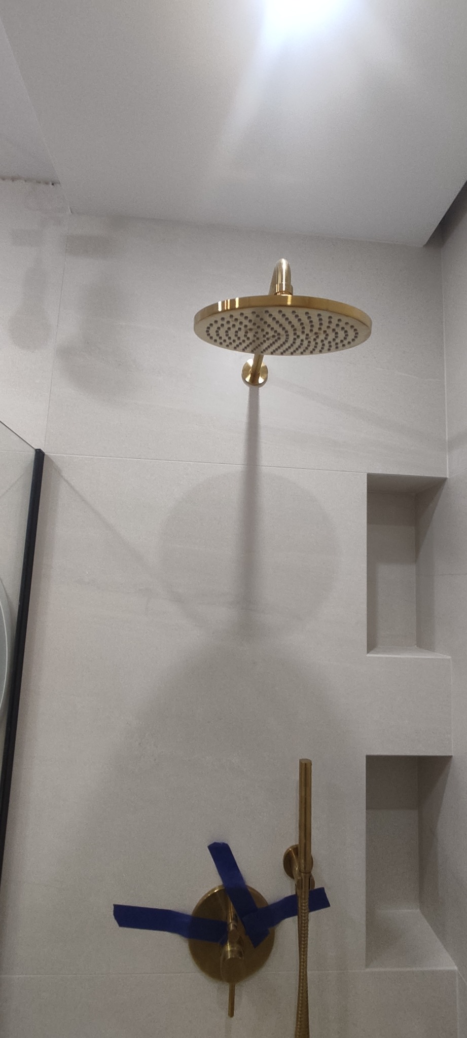 Złota deszczownica i bateria prysznicowa w trakcie montażu, zabezpieczone niebieską taśmą malarską na tle jasnych, kwadratowych płytek w nowoczesnej łazience.