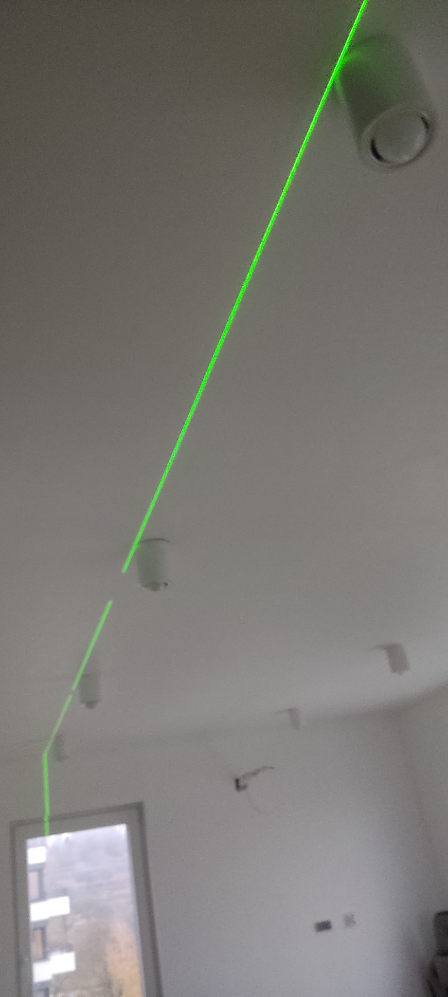 Laserowy poziom wyznacza linię na suficie z zamontowanymi punktowymi oprawami oświetleniowymi, widoczne okno w tle.