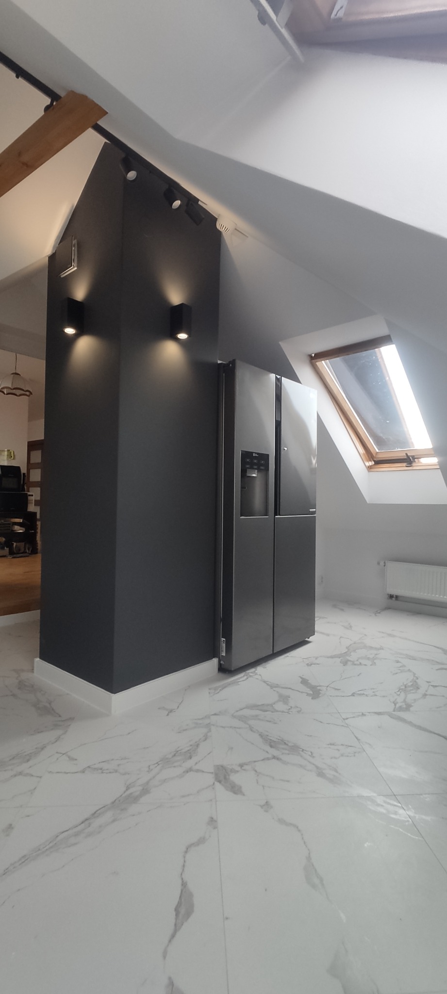 Szare wnętrze poddasza z marmurową podłogą, ciemną kolumną z oświetleniem punktowym i srebrną lodówką side-by-side przy ścianie ze skośnym oknem.