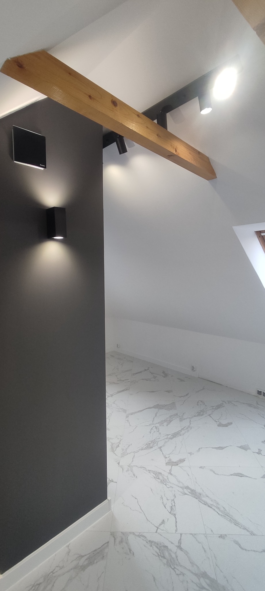 Szara ściana z czarnym kinkietem i czarnym głośnikiem, drewniana belka z reflektorami, marmurowe płytki podłogowe w nowo wykończonym pokoju na poddaszu.
