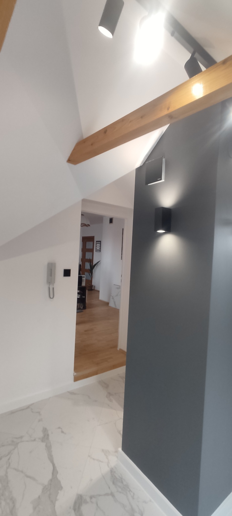 Nowoczesne wnętrze z szarą ścianą, oświetleniem punktowym, drewnianą belką stropową i podłogą z białego marmuru, widok na sąsiednie pomieszczenie z drewnianą podłogą.