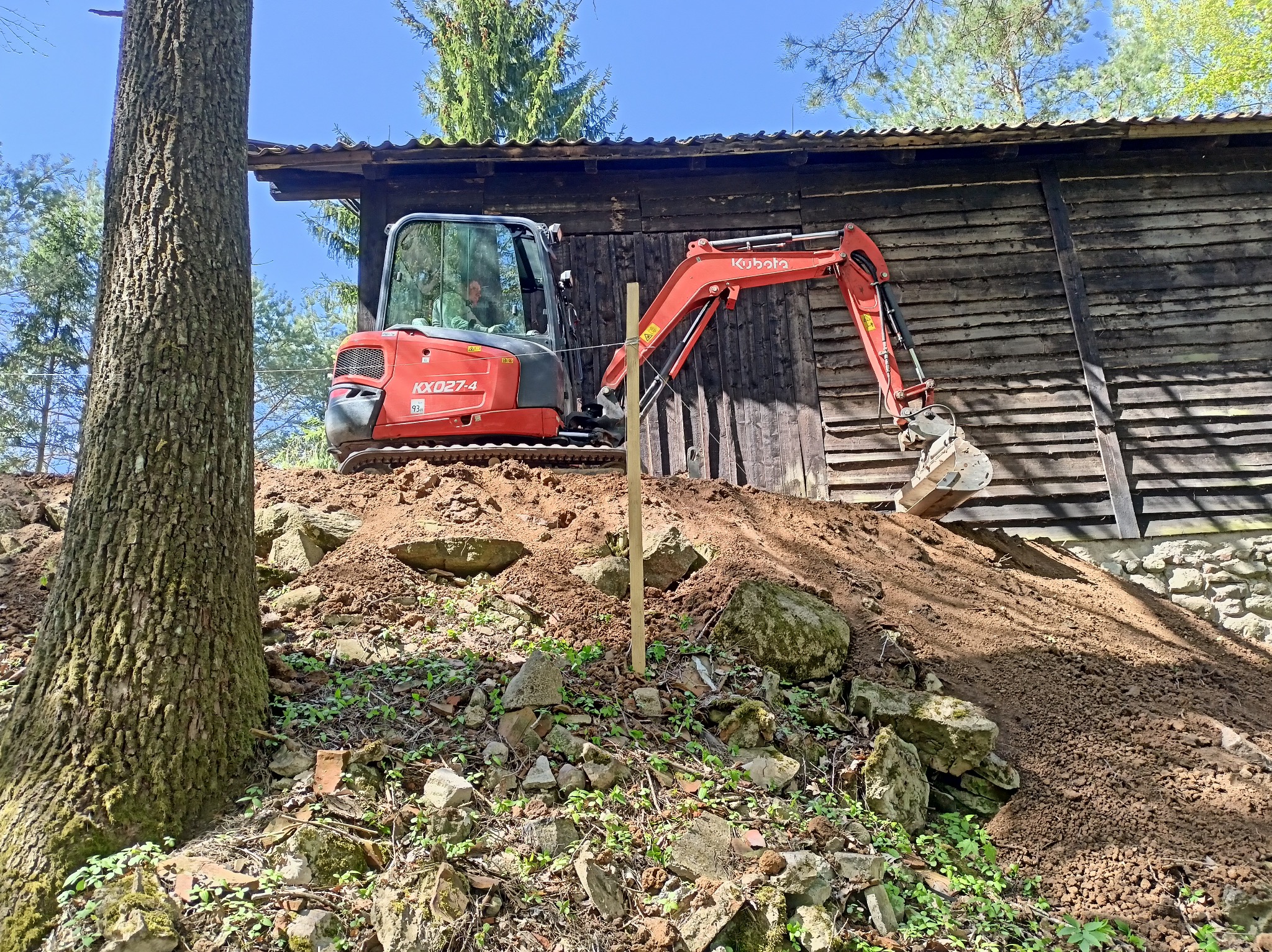 Czerwona minikoparka Kubota KX027-4 na nierównym terenie z ziemią i kamieniami, przy drewnianym budynku, widoczny słupek wyznaczający teren.