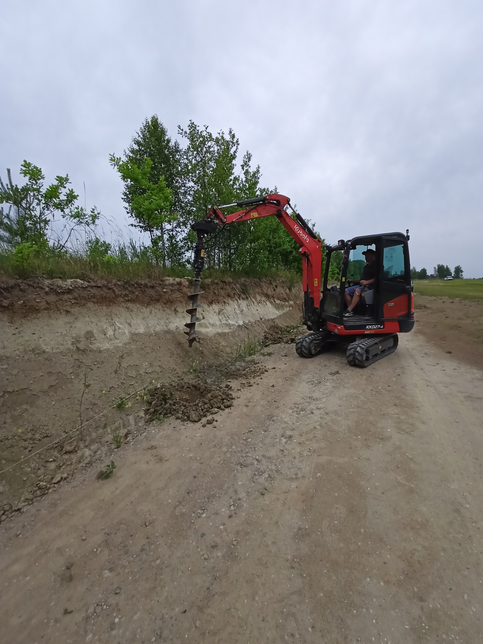 Czerwona mini koparka Kubota wierci otwór w ziemi wzdłuż wykopu na gruncie piaszczystym, operator w kabinie, drzewa w tle.