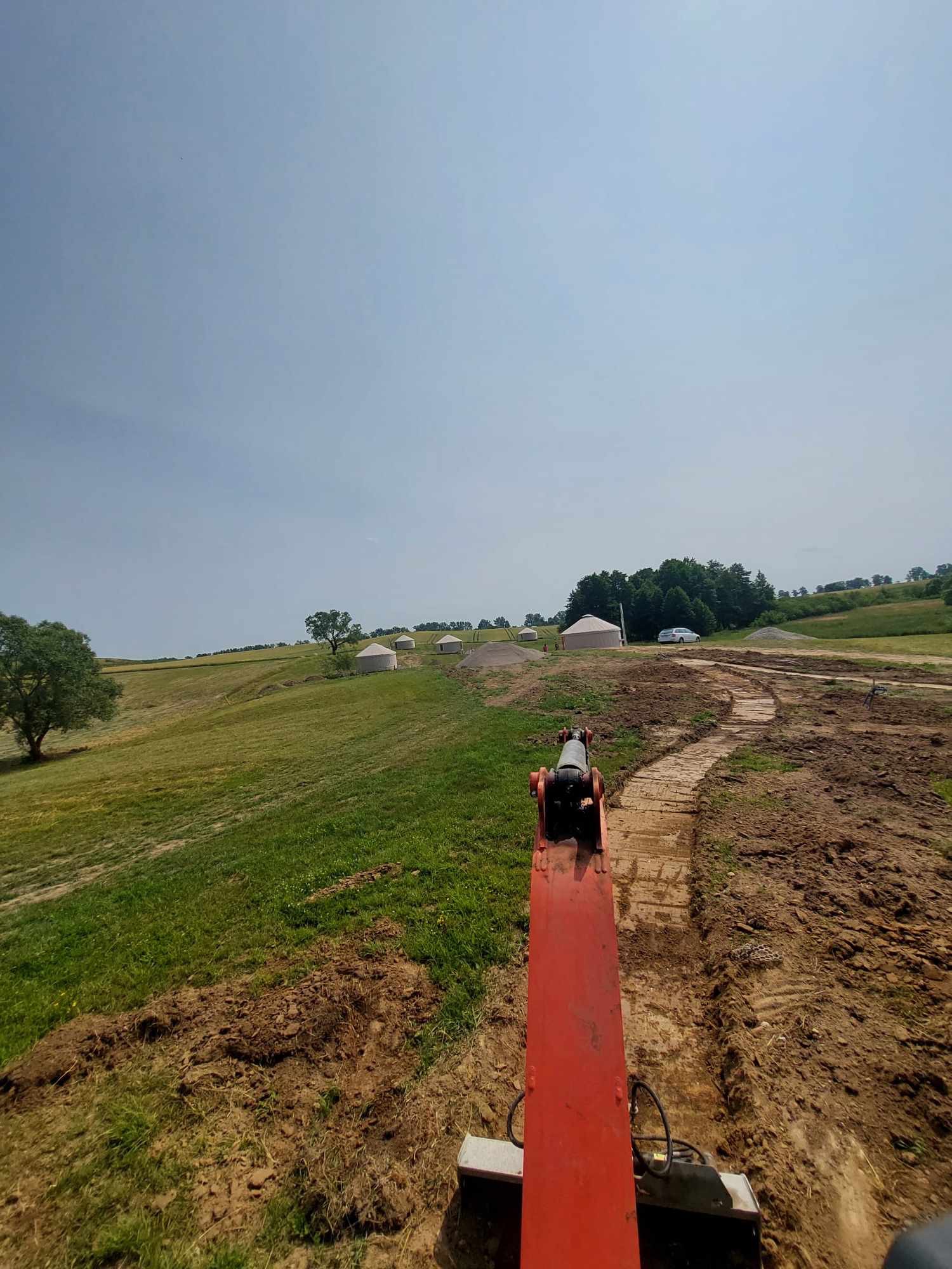 Widok z kabiny koparki na wykopany wąski rów na zielonym wzgórzu z kilkoma jurami w tle, w słoneczny dzień.