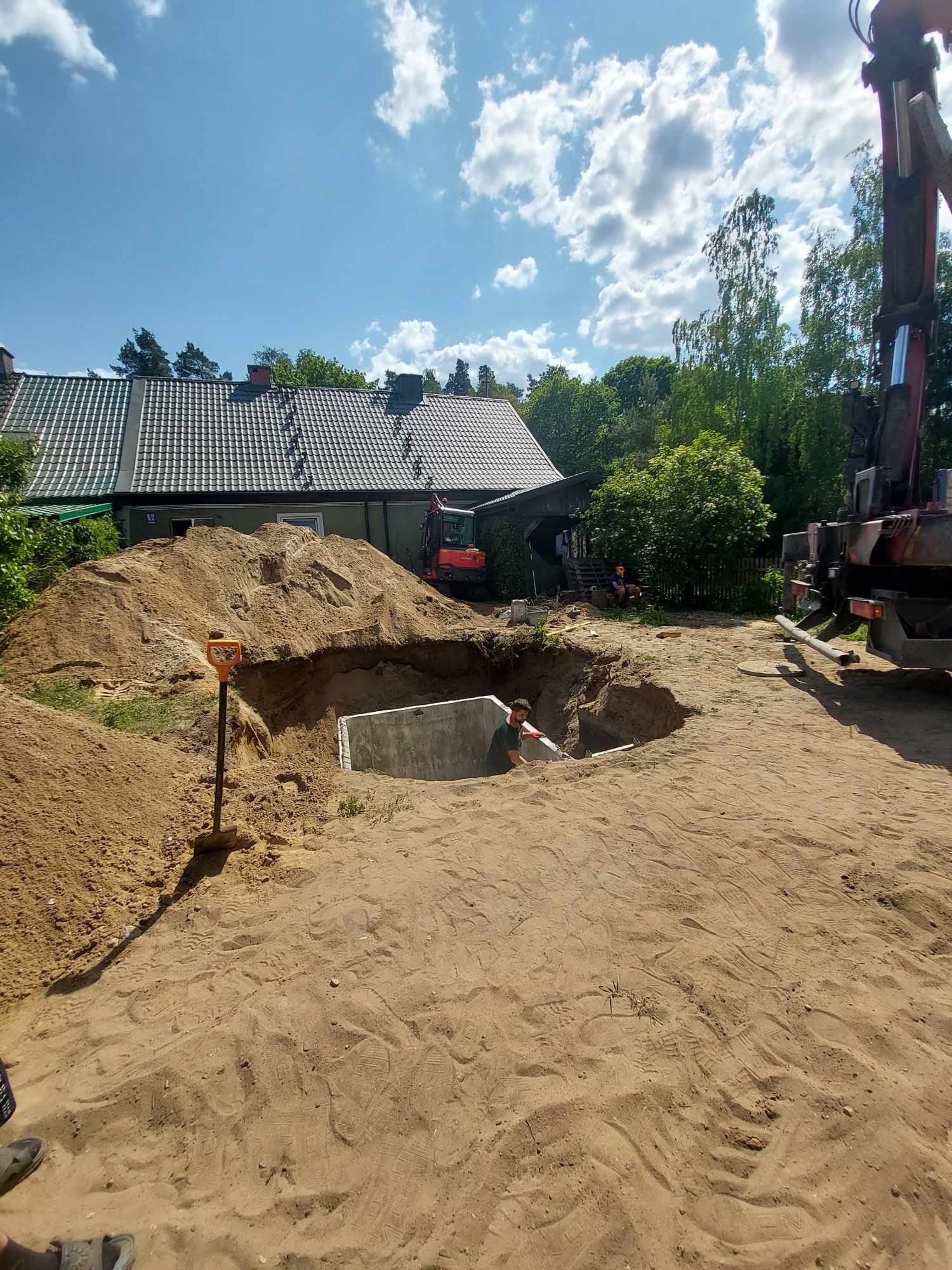 Prace ziemne w Mrągowie: Wykop z betonowymi ścianami, prawdopodobnie pod szambo, otoczony stertą piasku, z widocznym koparką w tle i dźwigiem po prawej stronie. W wykopie widoczna osoba...