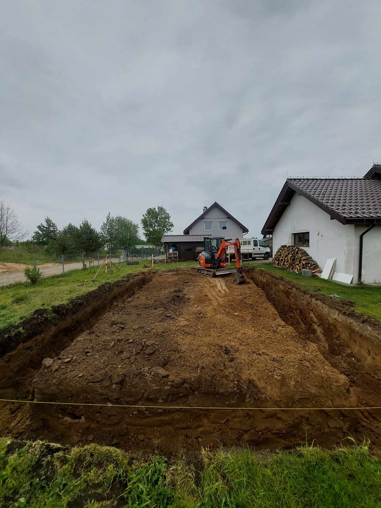 Prostokątny wykop w ziemi, z mini koparką w tle, widoczne domy w oddali i stos drewna przy jednym z nich. Grunt pod budowę przygotowany, wytyczony żółtą linką.