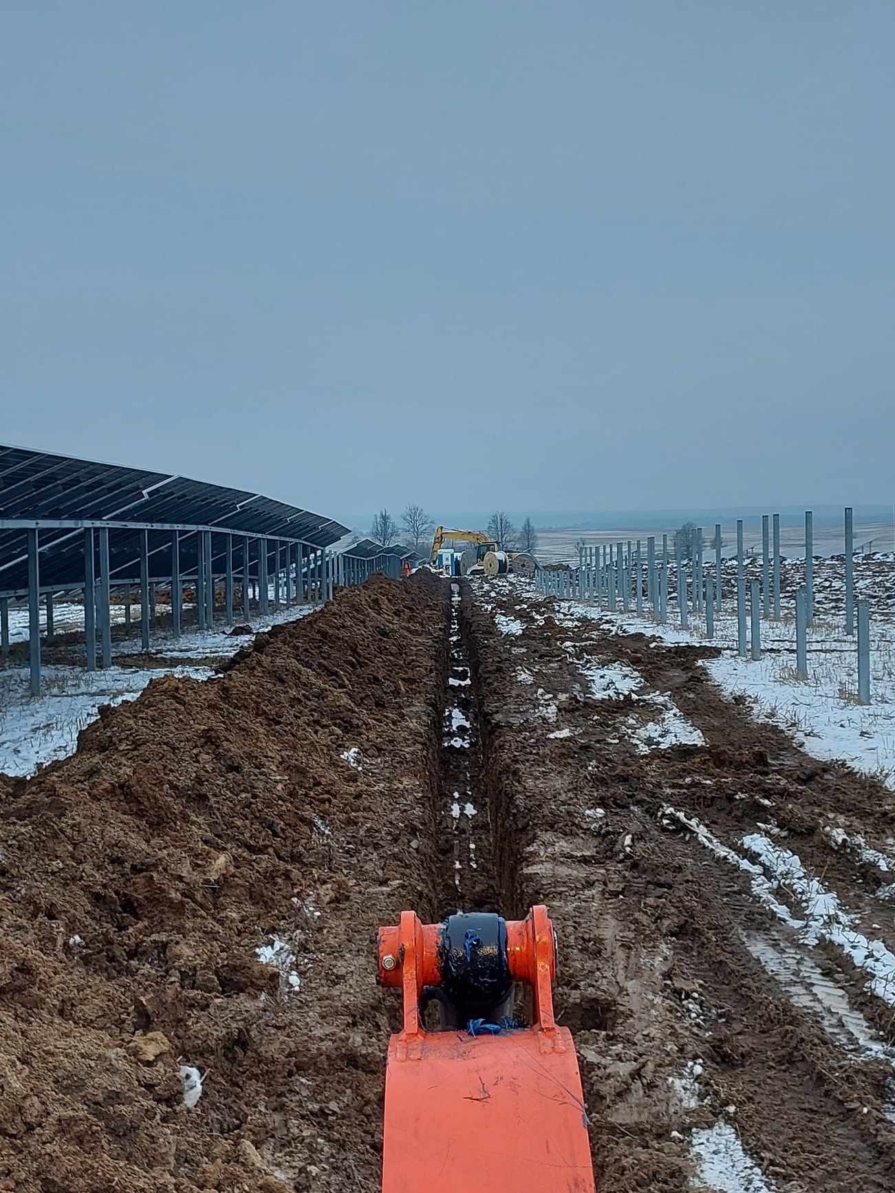 Wąski wykop pod instalacje na polu z panelami słonecznymi w tle, wykonany w zimowych warunkach, z widocznym fragmentem maszyny wykopującej.
