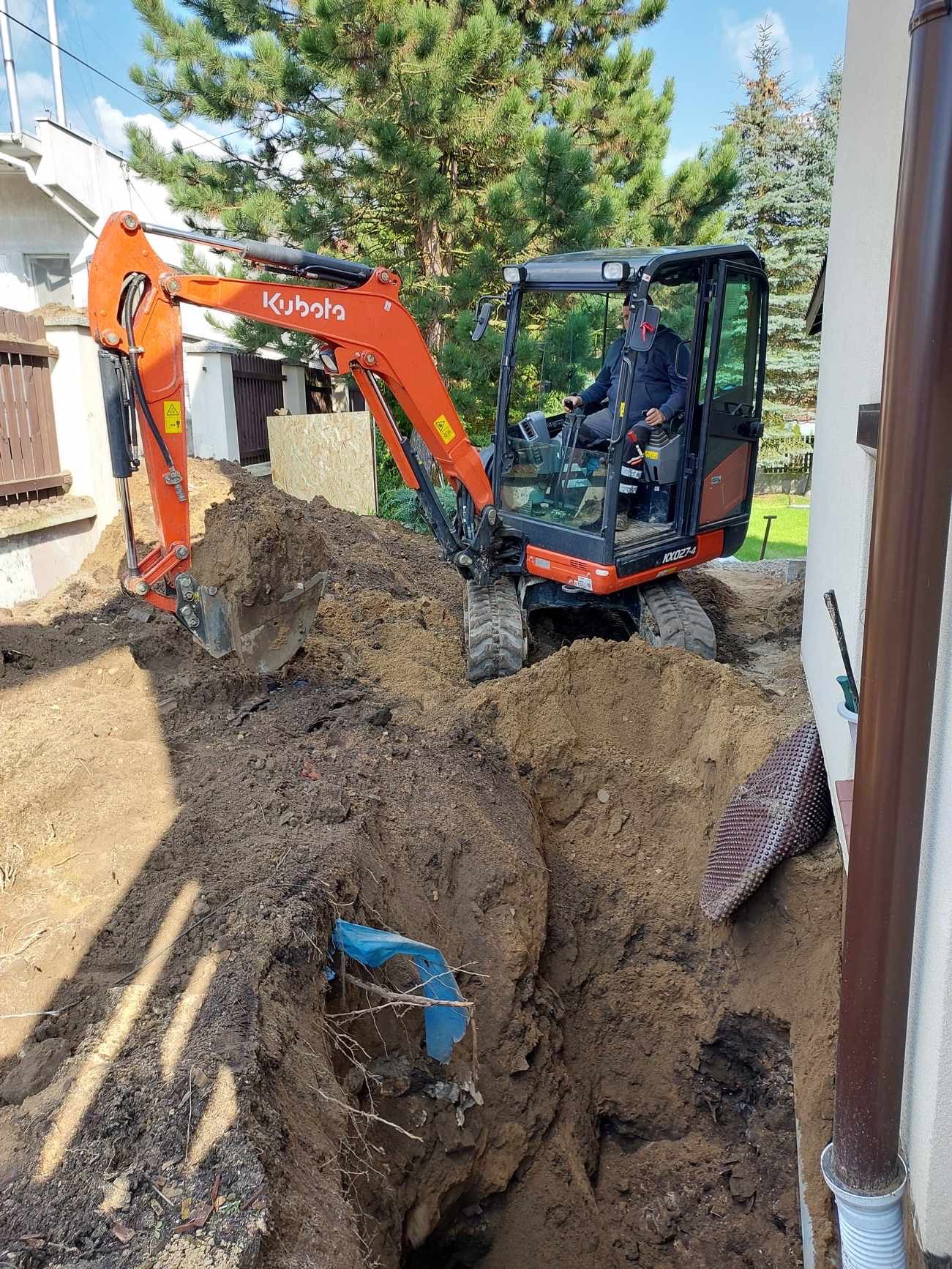 Pomarańczowa mini koparka Kubota w trakcie pracy przy wykopie wąskiego rowu w ziemi, widoczny niebieski fragment instalacji w wykopie, operator w kabinie, drzewa w tle.