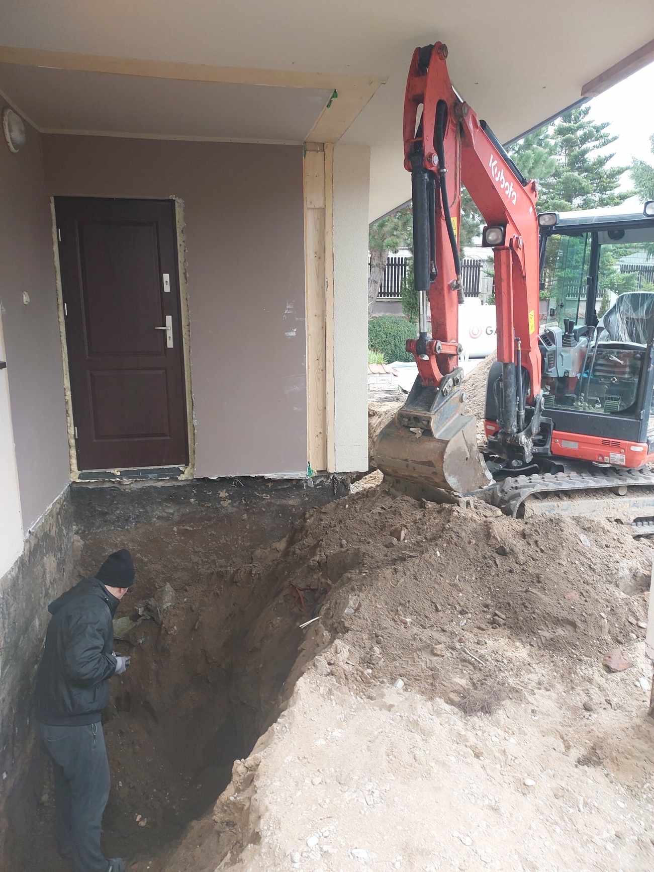 Czerwona mini koparka Kubota przy wykopie fundamentowym, obok stoi pracownik w czarnej kurtce i czapce, widoczny fragment budynku z brązowymi drzwiami.