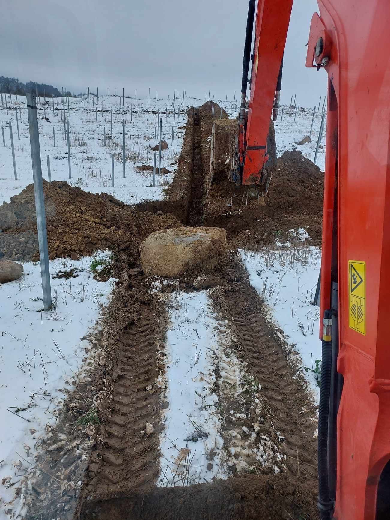 Pomarańczowa koparka w trakcie prac ziemnych na zaśnieżonym terenie, widoczny wykop i duży kamień w dole wykopu, w tle metalowe słupki.