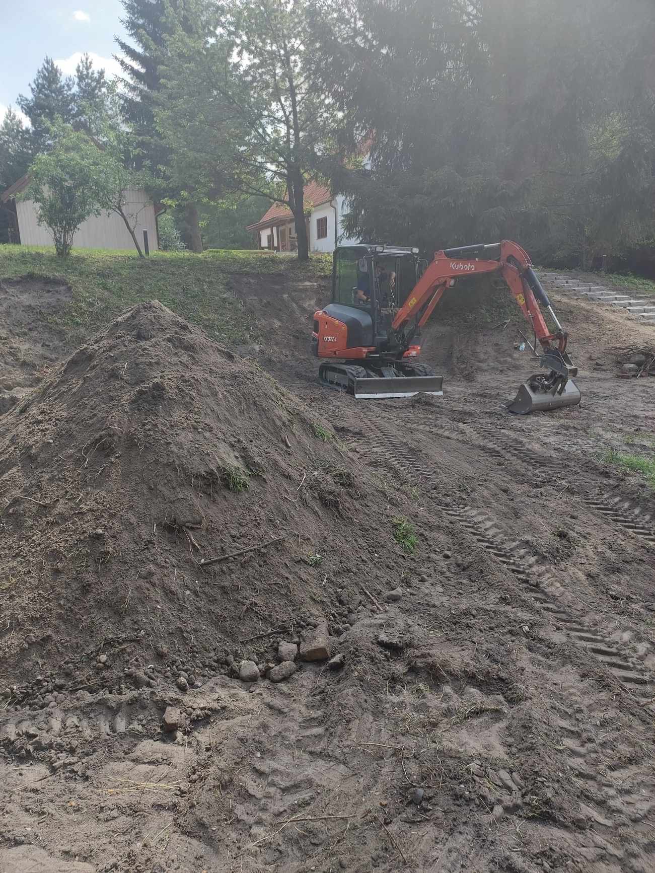 Pomarańczowa mini koparka Kubota na nierównym terenie z dużą stertą ziemi w tle, widoczne ślady opon, budynek w oddali.