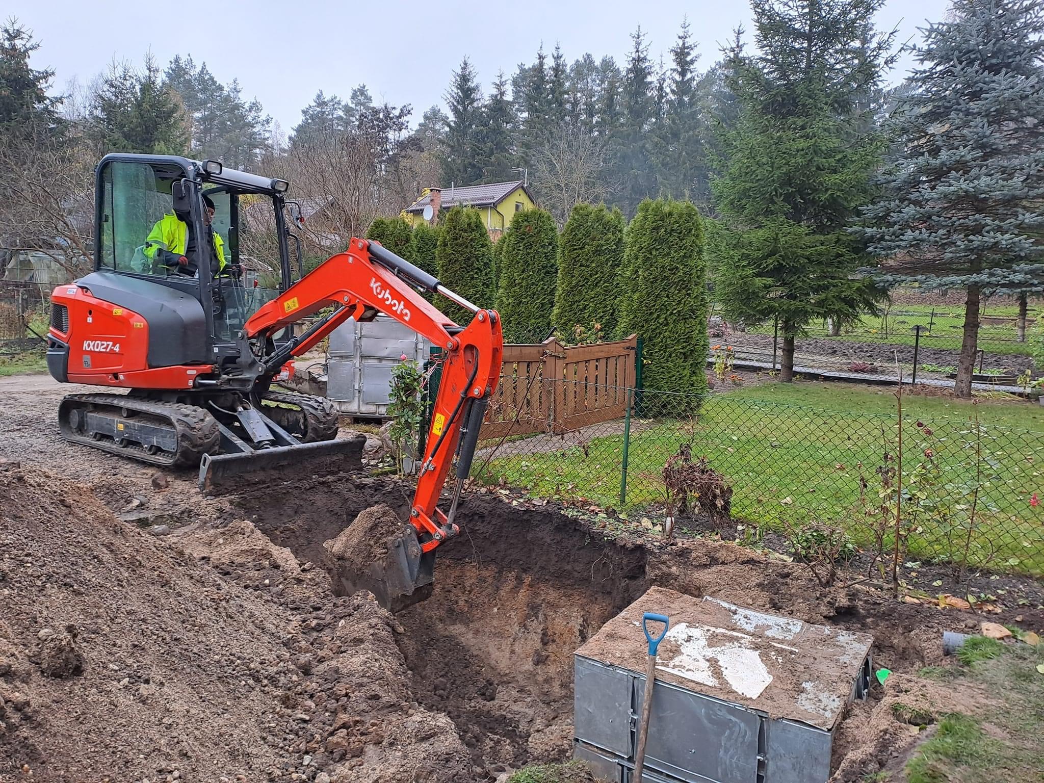 Pomarańczowa mini koparka Kubota KX027-4 w trakcie wykopu głębokiego rowu w ogrodzie, obok leżą elementy szamba betonowego, operator w żółtej kamizelce, pochmurny dzień.