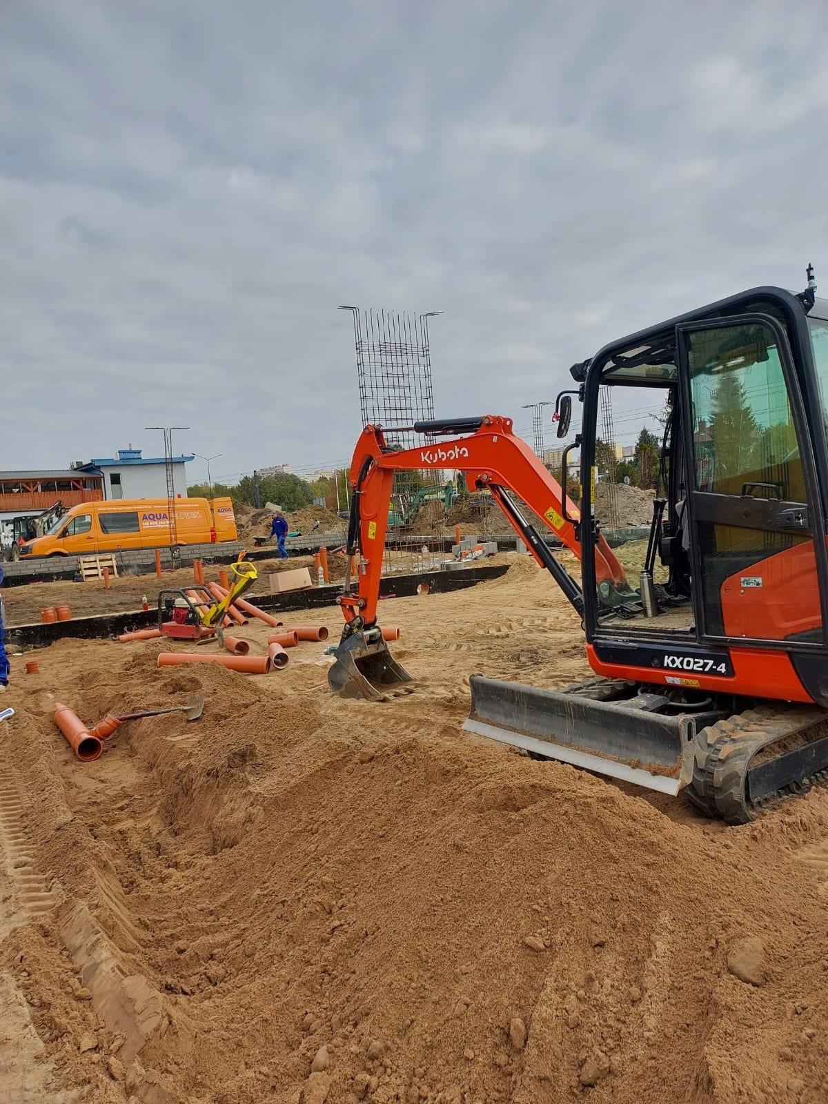 Pomarańczowa mini koparka Kubota KX027-4 na piaszczystym terenie budowy, widoczne rury kanalizacyjne, fragmenty fundamentów i zbrojenia, w tle żółty bus i pracownik w niebieskim kombinezonie.