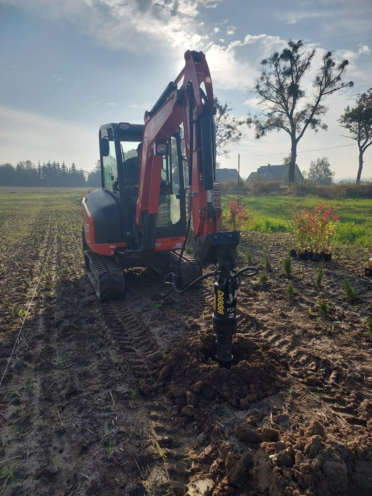 Mini koparka Kubota z wiertnicą hydrauliczną Digga PDX2 wiercąca otwór w grządkach, w tle drzewa i budynki pod jasnym niebem.
