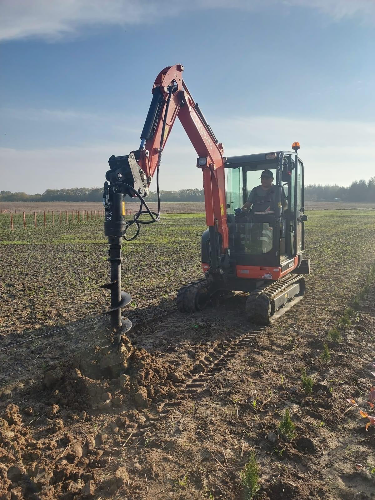Czerwona mini-koparka Kubota z wiertnicą hydrauliczną wiercąca otwór w polu uprawnym, widoczne ślady gąsienic i operator w kabinie.