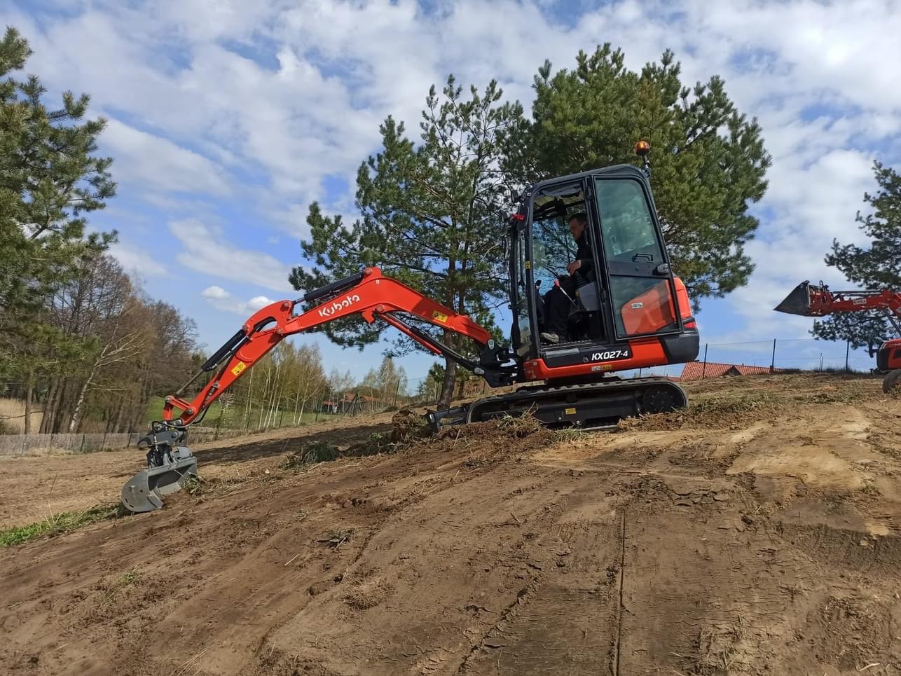 Pomarańczowa mini koparka Kubota KX027-4 na nierównym, piaszczystym terenie z widocznymi śladami pracy, operator w kabinie, w tle drzewa i błękitne niebo z chmurami.