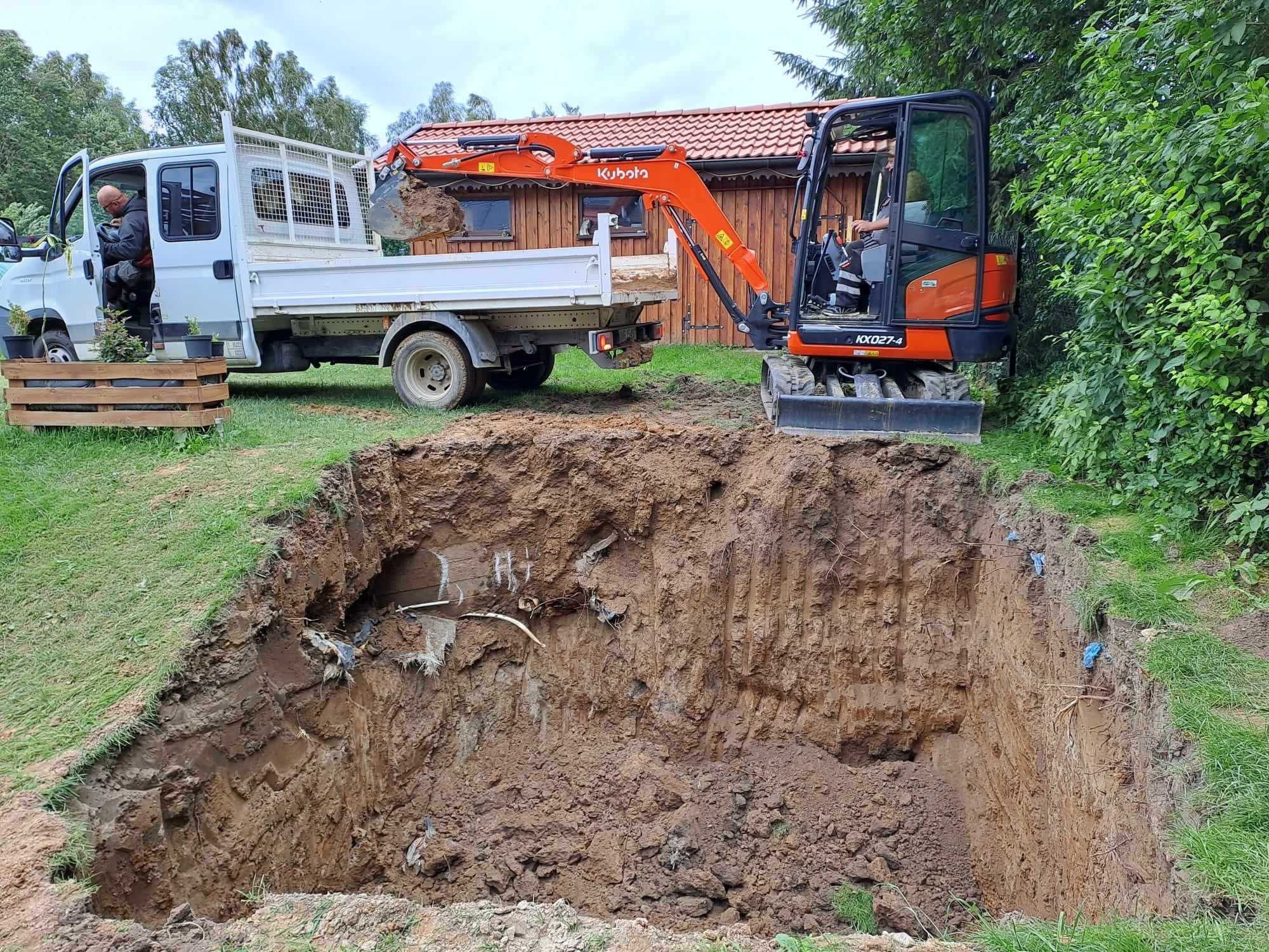 Pomarańczowa mini koparka Kubota nabiera ziemię z wykopu i wysypuje na skrzynię ładunkową białego samochodu dostawczego na tle drewnianego domku i zieleni.