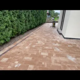 Nowa nawierzchnia z kostki brukowej w odcieniach brązu, ułożona wzdłuż żywopłotu i ściany budynku, z ubijarką wibracyjną i narzędziami w tle.