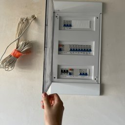 Twój Elektryk Tomek Usługi Elektryczne, Instalacje elektryczne, Awarie, Smart Home - Otwarta skrzynka elektryczna z bezpiecznikami na ścianie, widoczne kable. Ręka przytrzymuje drzwiczki skrzynki. Instalacja w trakcie prac wykończeniowych.