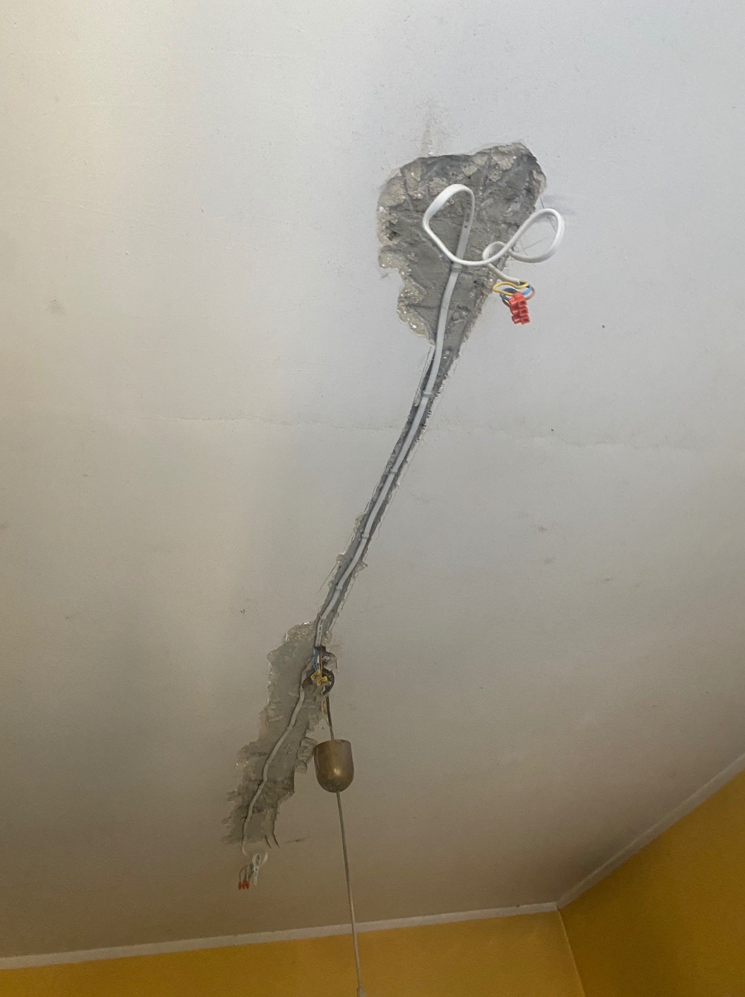 Uszkodzony sufit z widocznymi przewodami elektrycznymi i fragmentem lampy. Widoczne ślady po kuciu w suficie. Przewody w białej izolacji. Żółta ściana w tle.