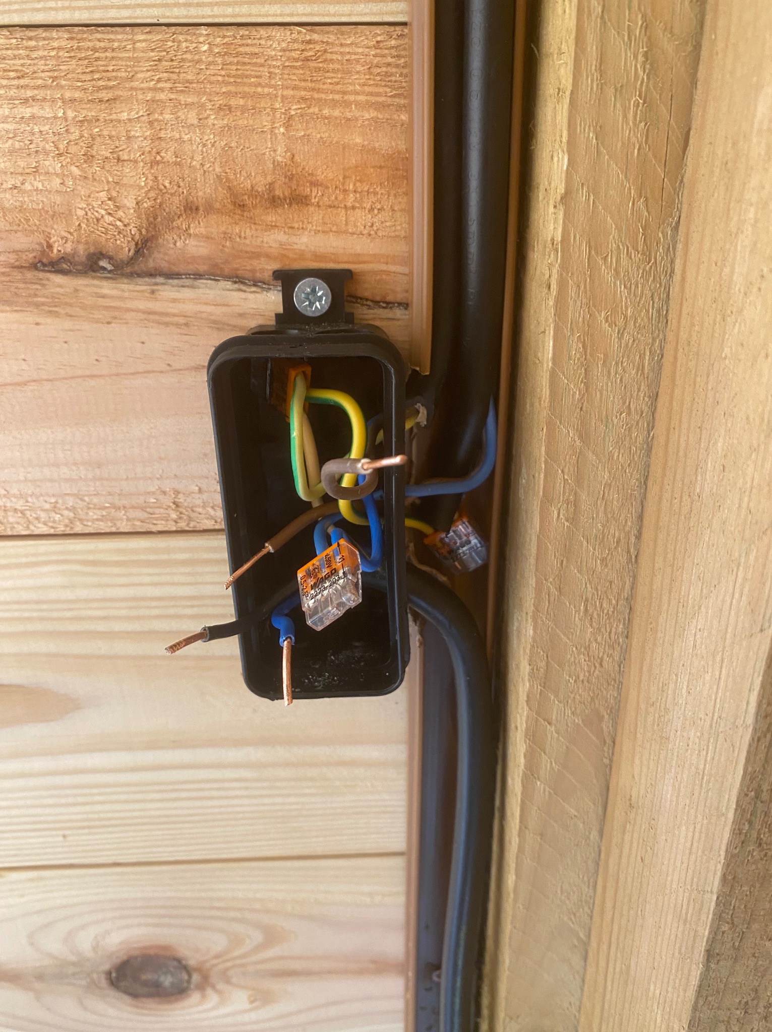 Otwarta puszka elektryczna z widocznymi przewodami i złączkami WAGO, zamontowana na drewnianej ścianie. Czarna rura instalacyjna biegnie wzdłuż ściany.