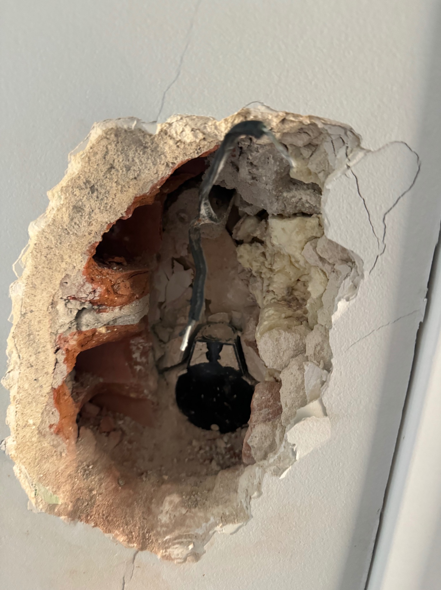 Otwór w ścianie z widocznymi przewodami elektrycznymi i fragmentami cegły, przygotowany do montażu oświetlenia. Widoczne pęknięcia na ścianie.