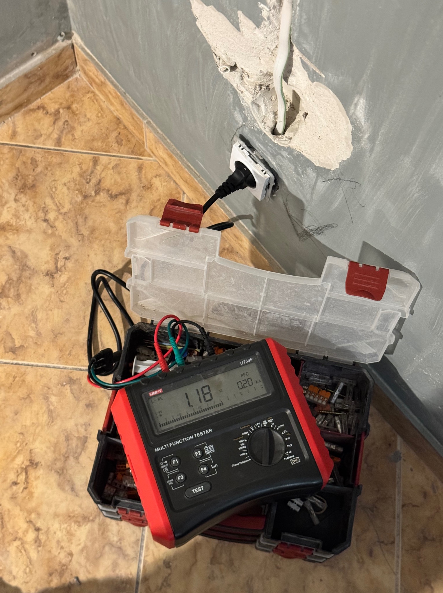 Pomiar napięcia w gniazdku elektrycznym za pomocą miernika wielofunkcyjnego UNI-T UT595. Widoczne przewody, gniazdko w ścianie i skrzynka z narzędziami.