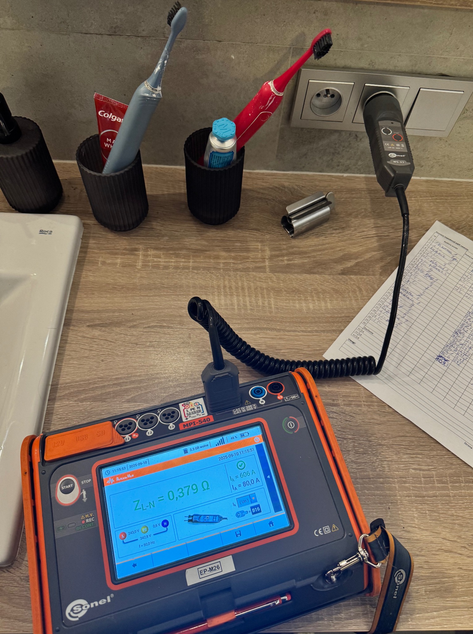 Pomiar impedancji pętli zwarcia w gniazdku elektrycznym w łazience, wykonany za pomocą miernika Sonel MPI-540. Widoczne gniazdko, przyrząd pomiarowy i notatki.