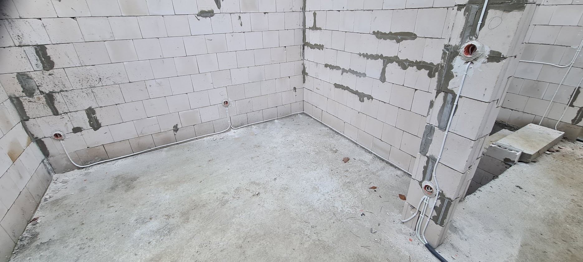 Surowy stan pomieszczenia z białych bloczków, widoczne poprowadzone kable elektryczne wzdłuż ścian i na słupie, puszki elektryczne zamontowane w ścianach.