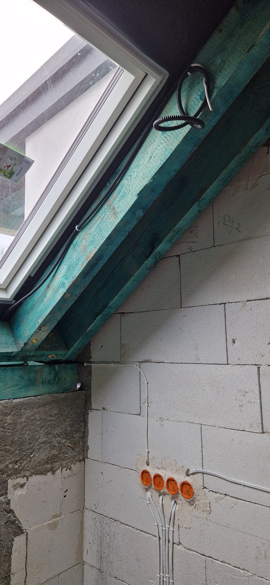 Montaż instalacji elektrycznej pod oknem dachowym w surowym stanie, widoczne kable w peszlu, puszki instalacyjne w ścianie z betonu komórkowego oraz drewniana konstrukcja okna pomalowana na zielono.