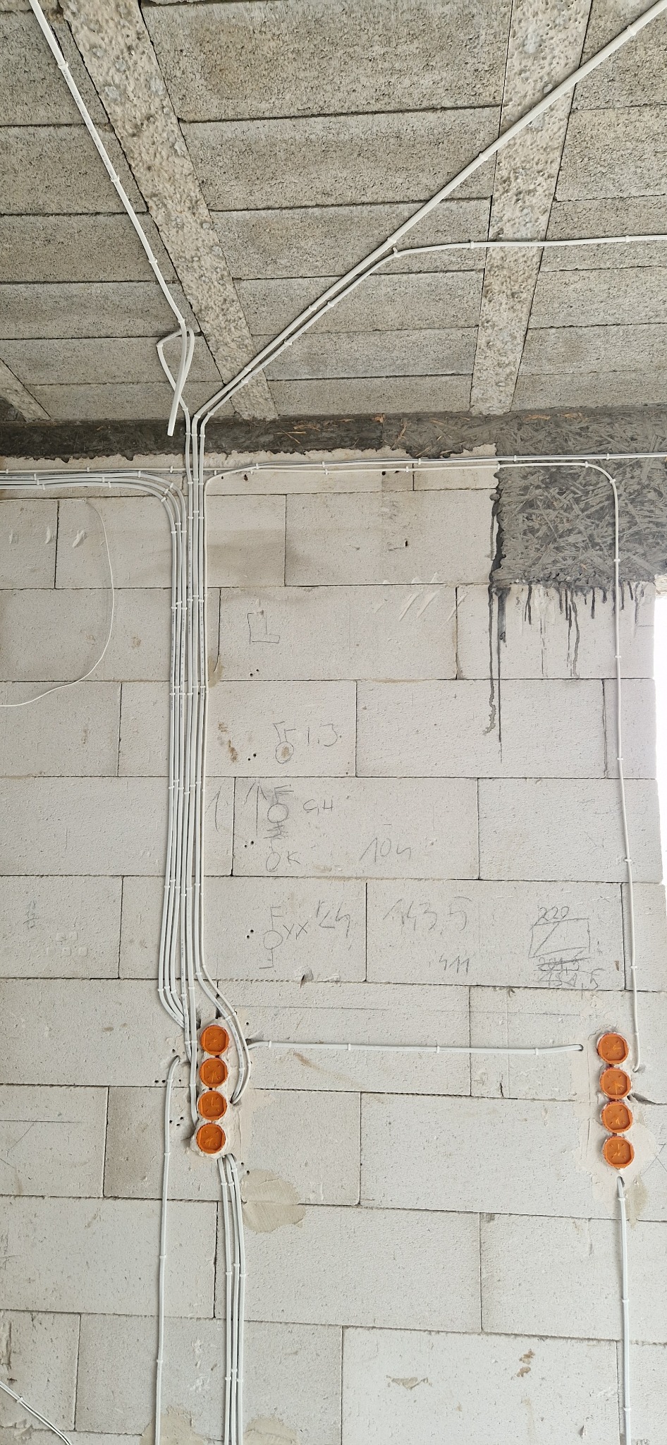Surowe wnętrze z instalacją elektryczną: białe przewody w peszlach prowadzone po bloczkach betonowych, z pomarańczowymi puszkami podtynkowymi i odręcznymi notatkami na ścianie.
