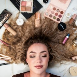 Fotograf Anna Sobula - Portret kobiety z zamkniętymi oczami otoczonej produktami do makijażu i pędzlami na marmurowym tle. Widoczne palety cieni, podkłady, pudry, błyszczyki i pędzle do makijażu.