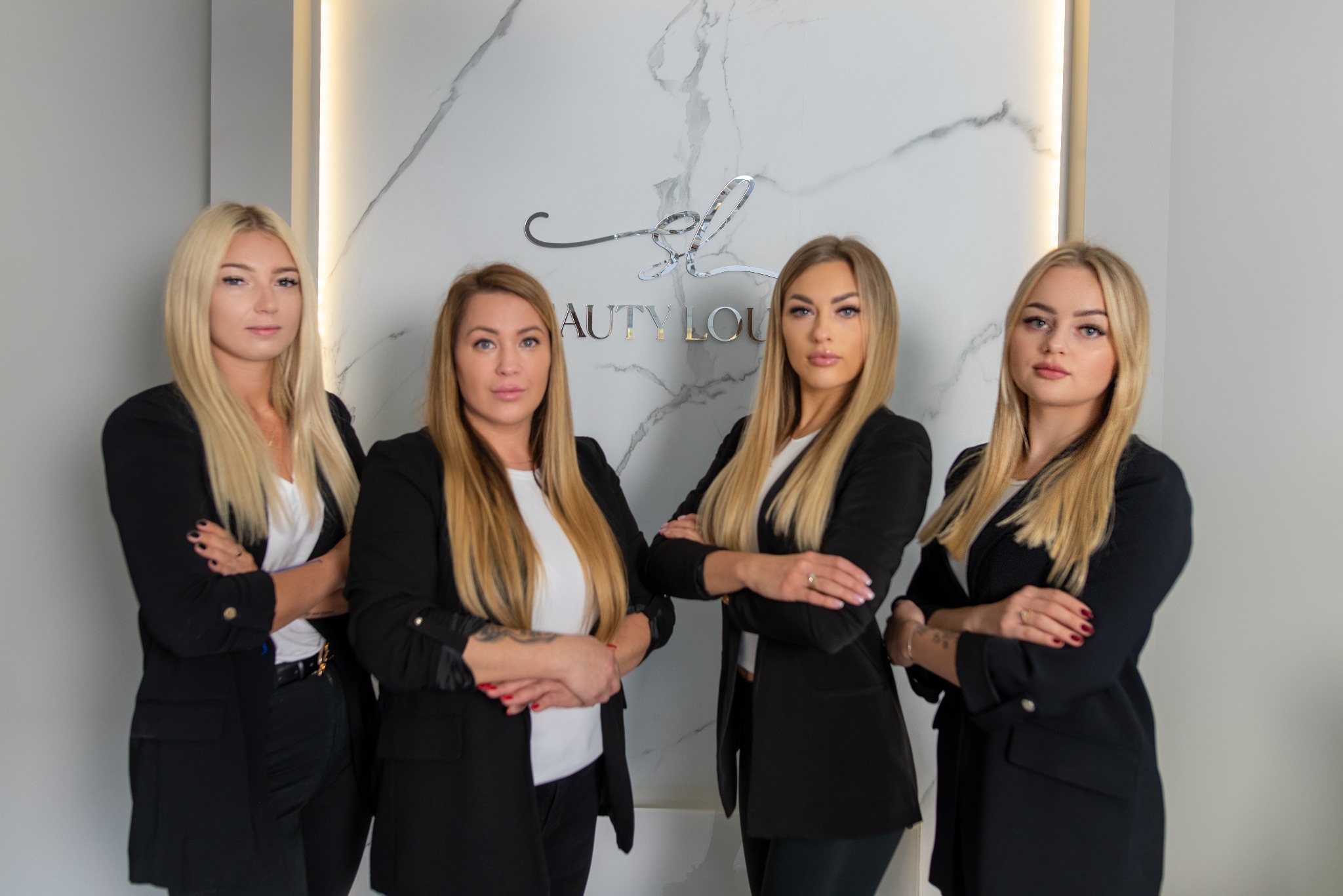 Portret grupowy czterech blondwłosych kobiet w czarnych marynarkach na tle marmurowej ściany z podświetlanym logo Beauty Lounge.