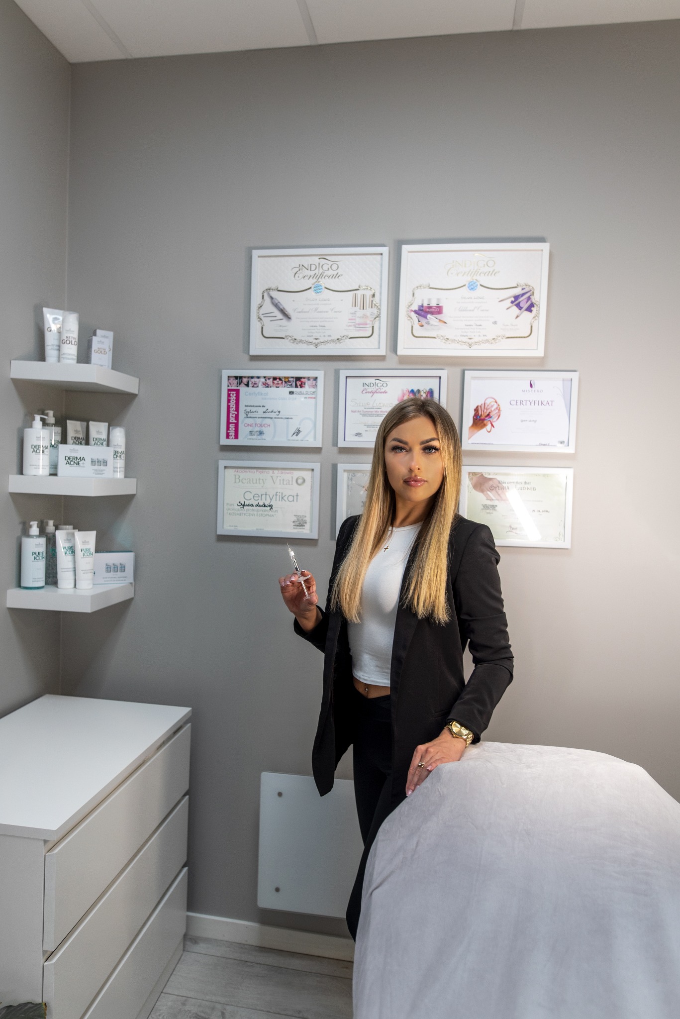 Kobieta w czarnym żakiecie trzyma strzykawkę w gabinecie kosmetycznym, na ścianie wiszą certyfikaty i półki z kosmetykami, obok znajduje się biała komoda i leżanka.