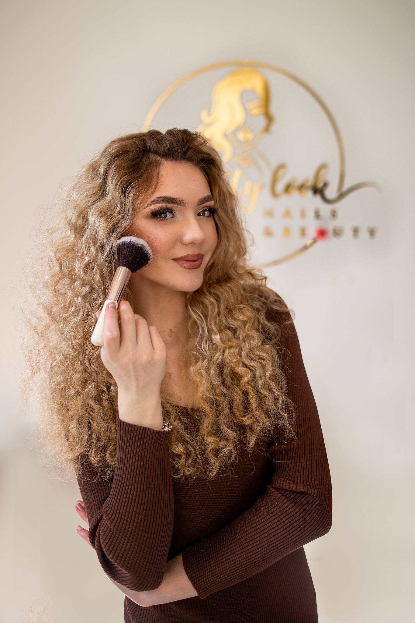 Portret kobiety z kręconymi blond włosami, trzymającej pędzel do makijażu przy policzku, z widocznym złotym logo salonu kosmetycznego w tle.