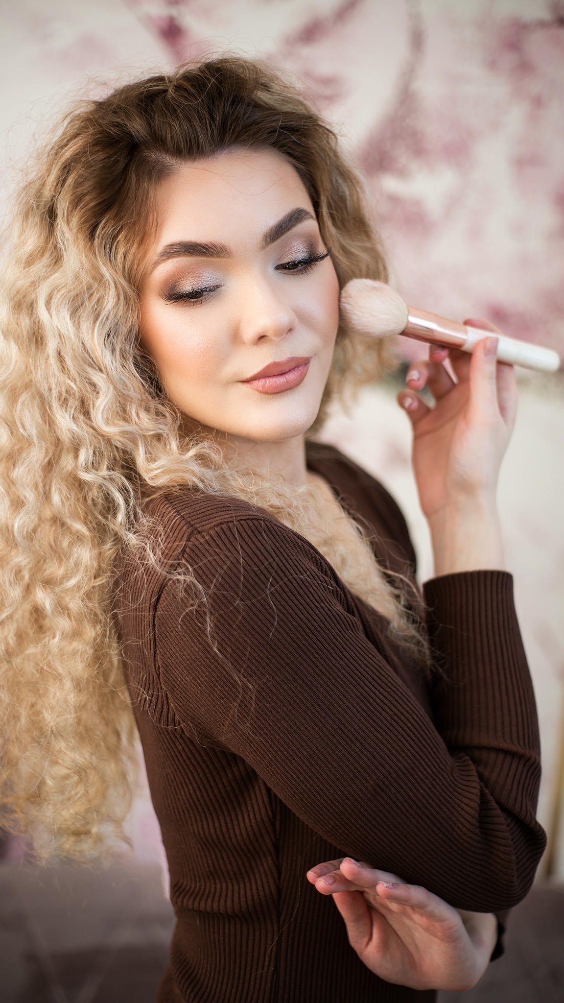 Portret kobiety z blond lokami, delikatny makijaż, aplikuje puder pędzlem na policzek, ubrana w brązowy sweter.
