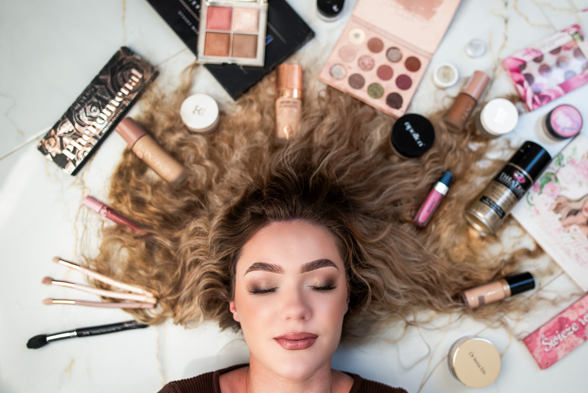 Portret kobiety z zamkniętymi oczami otoczonej produktami do makijażu i pędzlami na marmurowym tle. Widoczne palety cieni, podkłady, pudry, błyszczyki i pędzle do makijażu.