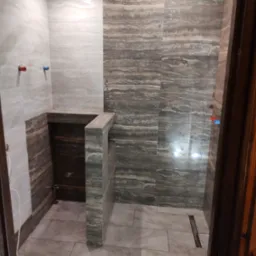 Wykończona łazienka z beżowo-brązowymi płytkami imitującymi trawertyn na ścianach i podłodze, widoczny odpływ liniowy i wystające rury instalacji wodnej.