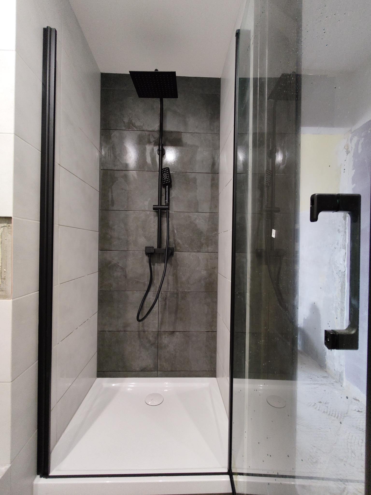 Nowoczesna kabina prysznicowa z czarną armaturą, szklanymi drzwiami i płytkami w odcieniach szarości, widoczne ślady prac wykończeniowych na ścianie zewnętrznej.