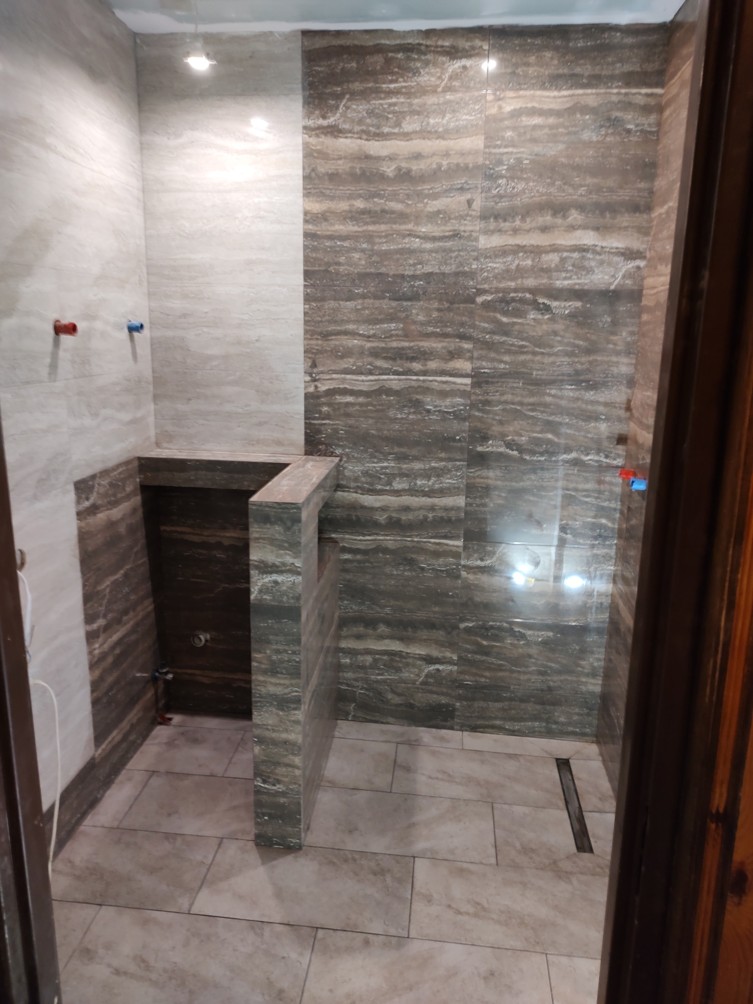 Wykończona łazienka z beżowo-brązowymi płytkami imitującymi trawertyn na ścianach i podłodze, widoczny odpływ liniowy i wystające rury instalacji wodnej.