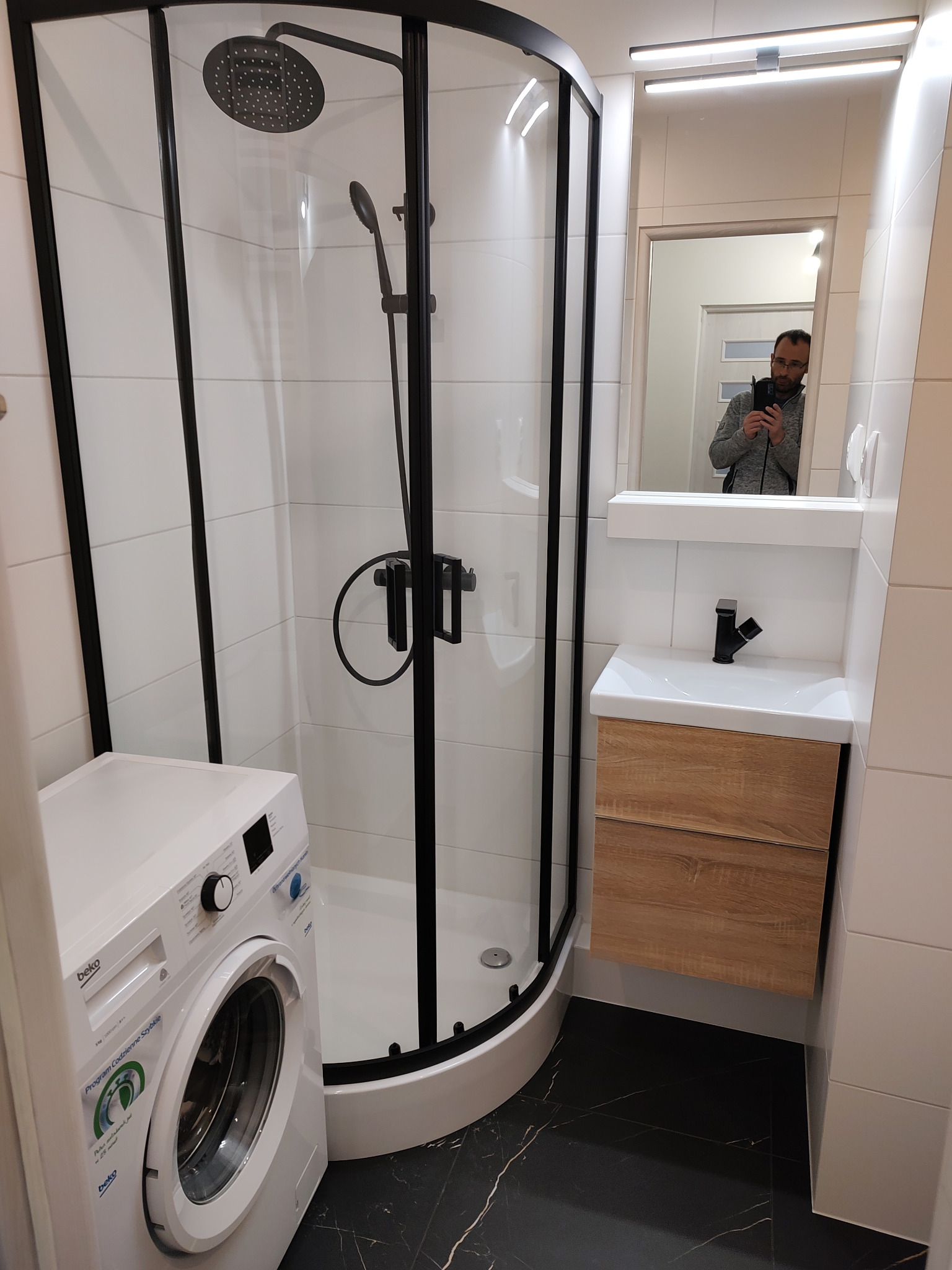 Nowoczesna łazienka z kabiną prysznicową z czarnymi profilami, umywalką z szafką w kolorze drewna i pralką Beko, widoczny fragment lustra z odbiciem osoby robiącej zdjęcie.