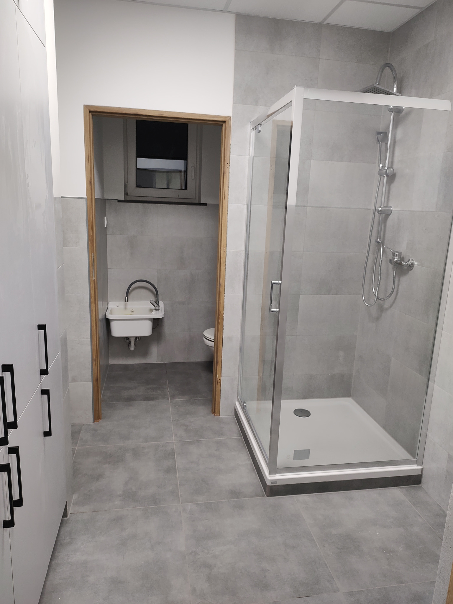 Nowoczesna łazienka z szarymi płytkami, kabiną prysznicową z hydromasażem i białą szafką z czarnymi uchwytami, widziana z perspektywy drzwi do drugiego pomieszczenia z umywalką.