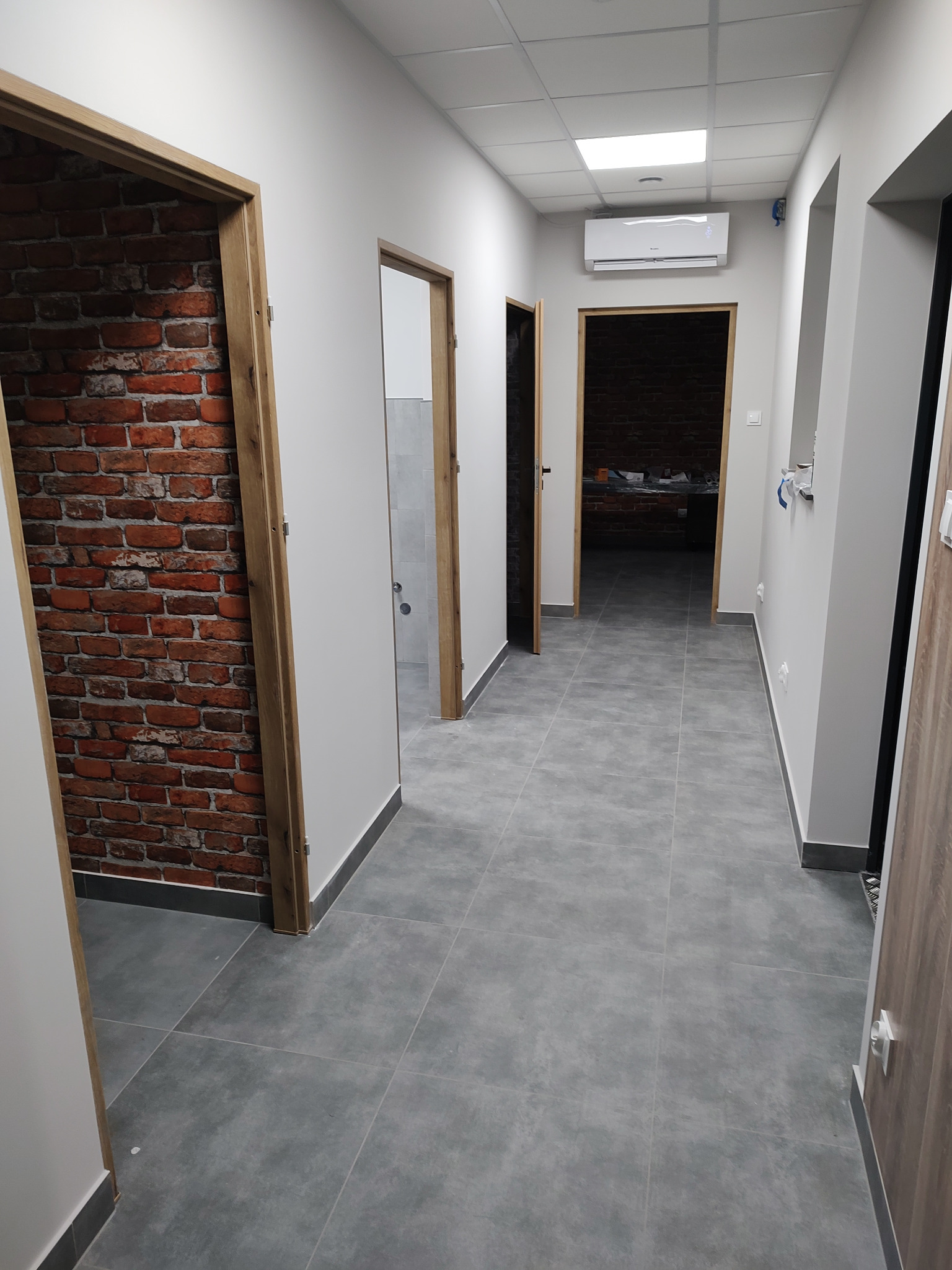 Korytarz biurowy po remoncie z szarymi płytkami podłogowymi, białymi ścianami, klimatyzacją i dekoracyjną ścianą z cegły.