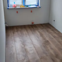 Ułożone panele podłogowe w pustym pomieszczeniu z oknem zabezpieczonym folią malarską i niepodłączonymi rurami instalacji wodnej.