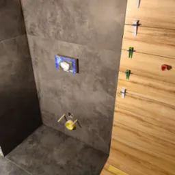 Częściowo wykończona łazienka z szarymi płytkami na ścianach i podłodze, widoczna instalacja podtynkowego WC oraz drewnopodobne płytki na ścianie z klipsami poziomującymi.