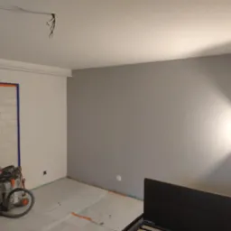 Pokój w trakcie malowania na szaro, zabezpieczona folią podłoga, widoczny odkurzacz przemysłowy i narzędzia, fragment łóżka w prawym dolnym rogu.