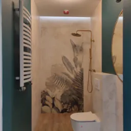 Nowoczesna łazienka z motywem tropikalnych liści na ścianie, złota armatura prysznicowa, podwieszana toaleta i grzejnik drabinkowy.