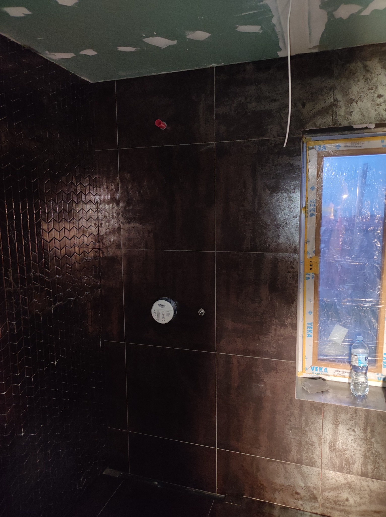 Ciemne, błyszczące płytki w trakcie montażu w łazience, widoczny fragment sufitu z niedokończonymi gładziami, zabezpieczone okno z butelką wody na parapecie, fragment ściany z dekoracyjnymi...