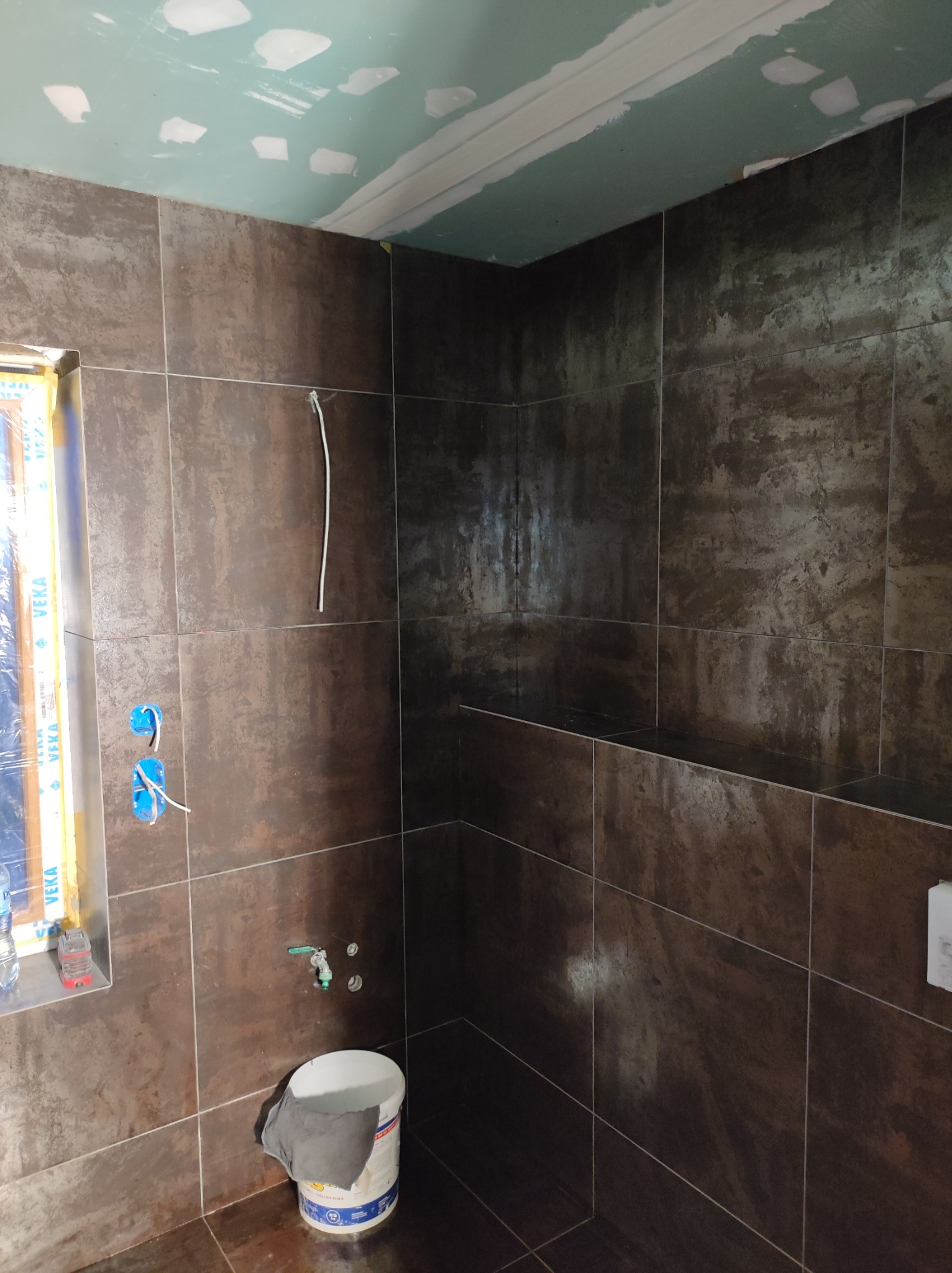 Brązowe płytki imitujące rdzę na ścianach łazienki w trakcie remontu, widoczne niezamaskowane okno z profilem VEKA, puszki elektryczne bez osprzętu, fragment sufitu z płyt gipsowo-kartonowych...