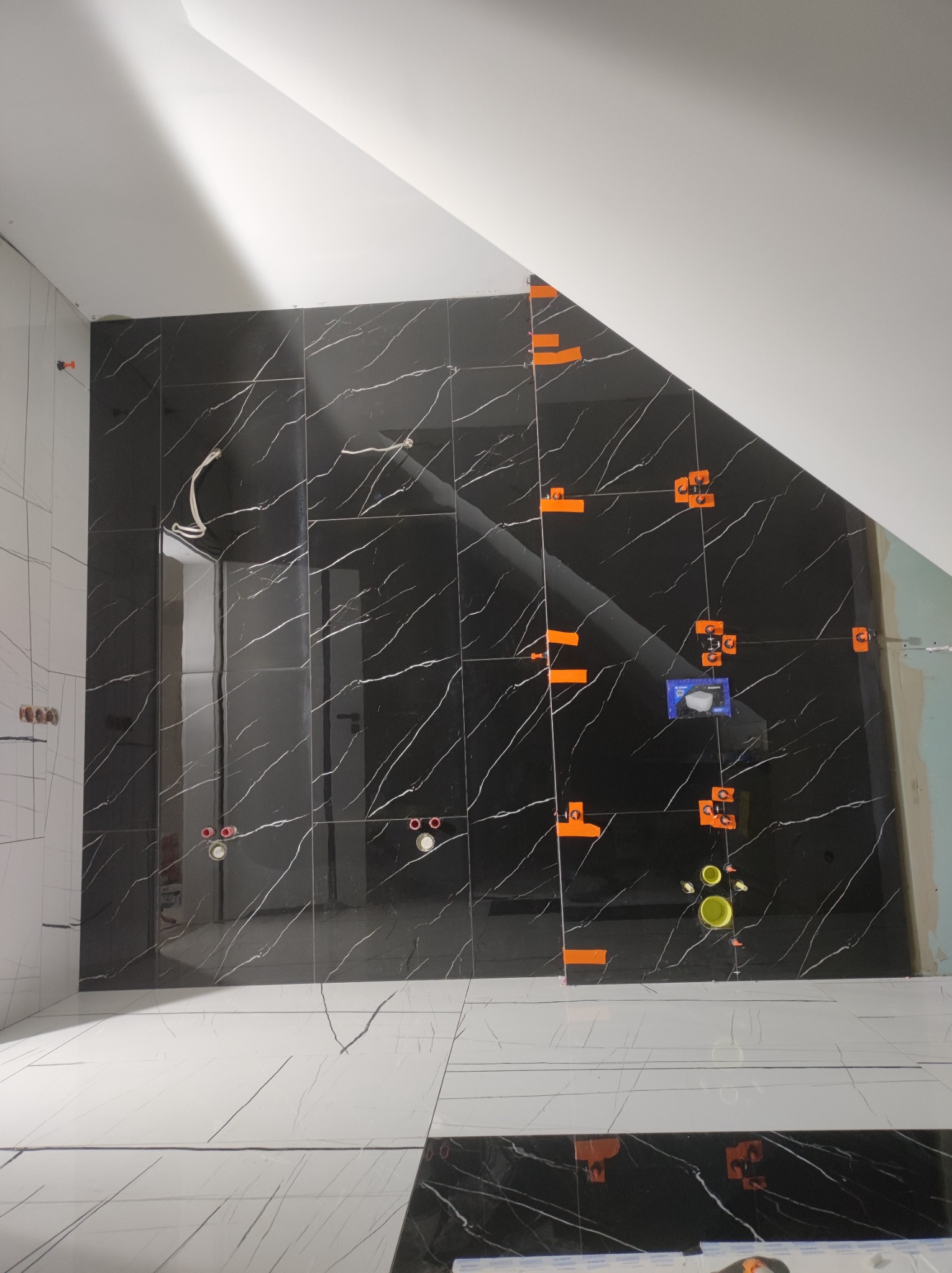Ściana i podłoga w trakcie układania czarno-białych płytek imitujących marmur, z widocznymi pomarańczowymi krzyżykami dystansowymi i elementami instalacji sanitarnej.